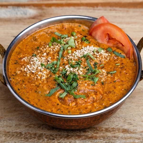 Vibrant orange curry in a metal serving bowl, topped with chopped nuts, fresh herbs, and tomato.