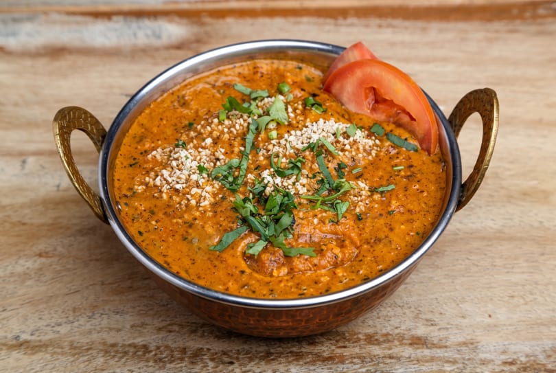 Vibrant orange curry in a metal serving bowl, topped with chopped nuts, fresh herbs, and tomato.