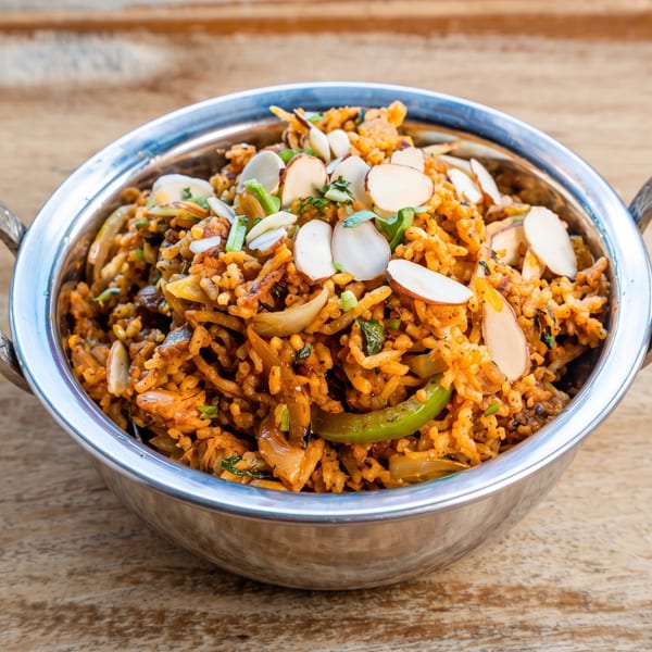 Spiced rice dish with onions, green peppers, and sliced almonds in a silver bowl on wood.
