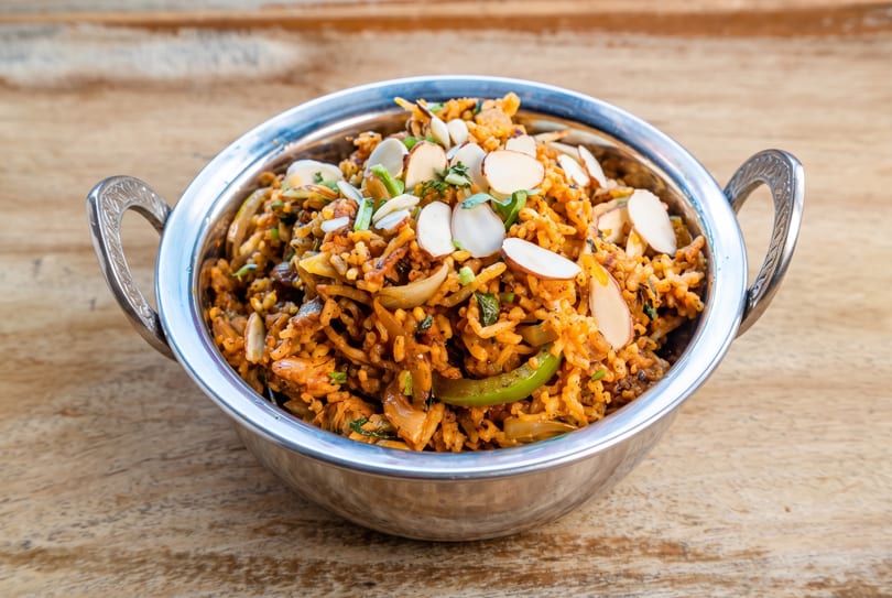 Spiced rice dish with onions, green peppers, and sliced almonds in a silver bowl on wood.