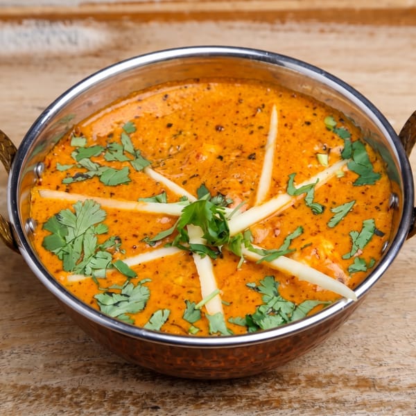Vibrant orange Indian curry in a traditional metal karahi, garnished with cilantro and ginger on wood.