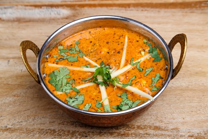 Vibrant orange Indian curry in a traditional metal karahi, garnished with cilantro and ginger on wood.