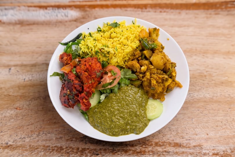 Colorful Indian thali with yellow rice, red chicken, green chutney, and potato curry on wood.