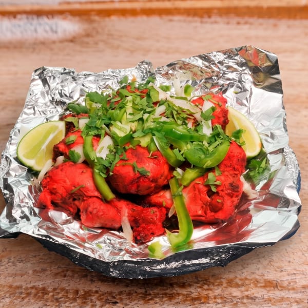 Bright red chicken pieces in foil, topped with green peppers, onions, cilantro, and lime wedges.