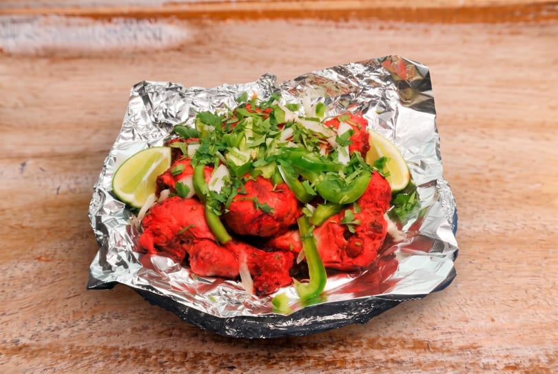 Bright red chicken pieces in foil, topped with green peppers, onions, cilantro, and lime wedges.