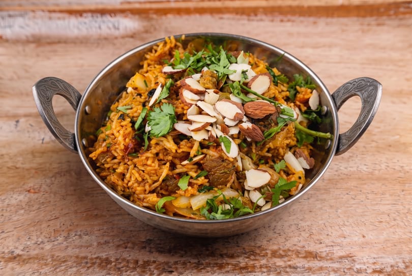 Spiced rice dish with meat, cilantro, and almonds in a decorative metal bowl on a wooden surface.
