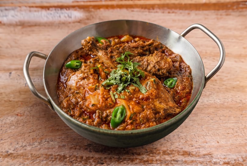 Chicken curry in a metal serving bowl, garnished with fresh cilantro and green chilies on a wooden table.