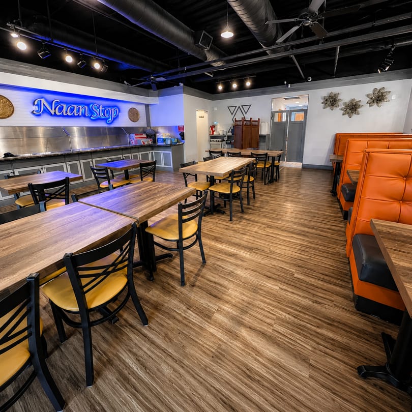 Restaurant interior with wooden tables, yellow chairs, orange booths, and a "Naan Stop" buffet line.